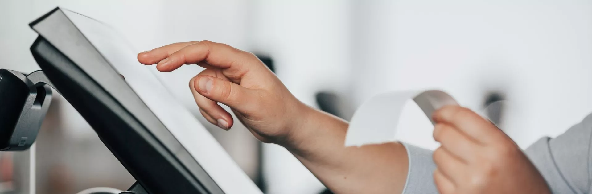 Mão segurando um cupom fiscal enquanto outra toca a tela de um terminal de atendimento com impressora acoplada, em ambiente de balcão.